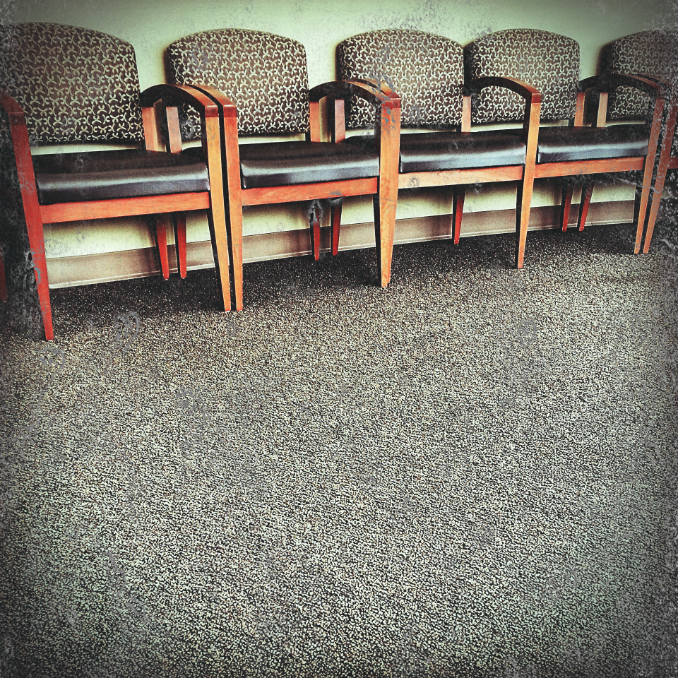 A colour photograph of wooden chairs placed arm-to-arm against a waiting room wall. Image by Alicia from Pixabay. Modified.