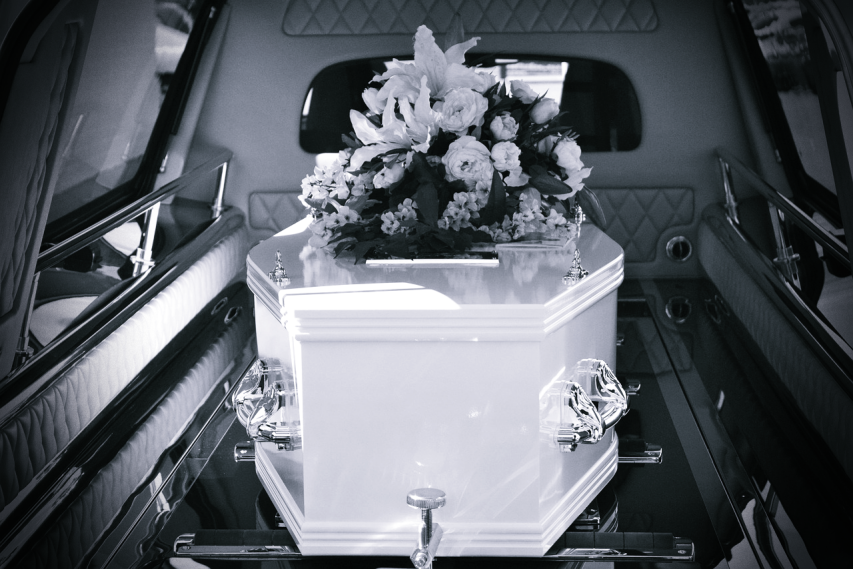 A monochrome photo of a coffin in the rear of a hearse, with an arrangement of flowers on top. Image by Carolyn Booth from Pixabay (modified).